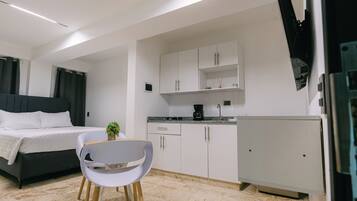 A modern bedroom with a bed, a small dining area with a chair, and a kitchenette with cabinets and a coffee maker.