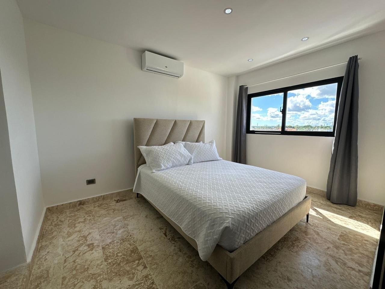 Un dormitorio con cama, ventana con cortinas, unidad de aire acondicionado y vista al cielo.
