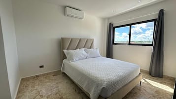A bedroom with a bed, a window with curtains, an air conditioning unit, and a view of the sky.
