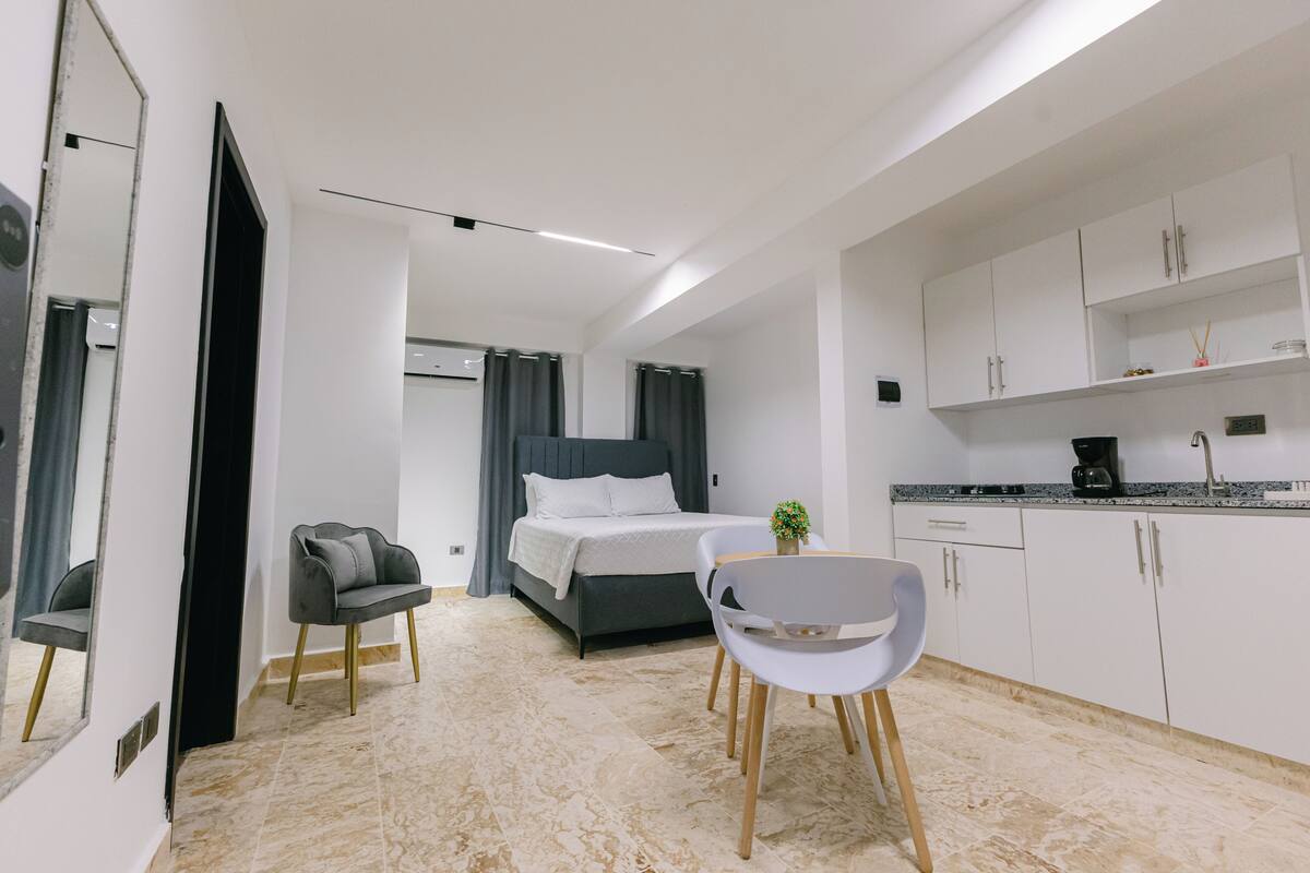 A modern hotel room with a bed, a desk, a chair, a small table, and a kitchen area.