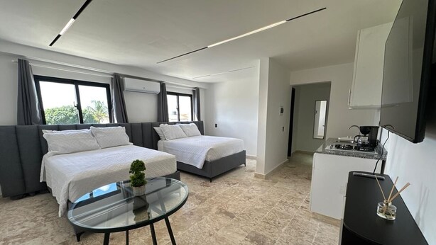 Et moderne hotelværelse med to senge, et glasbord, en lille plante, et tekøkken og et stort vindue.