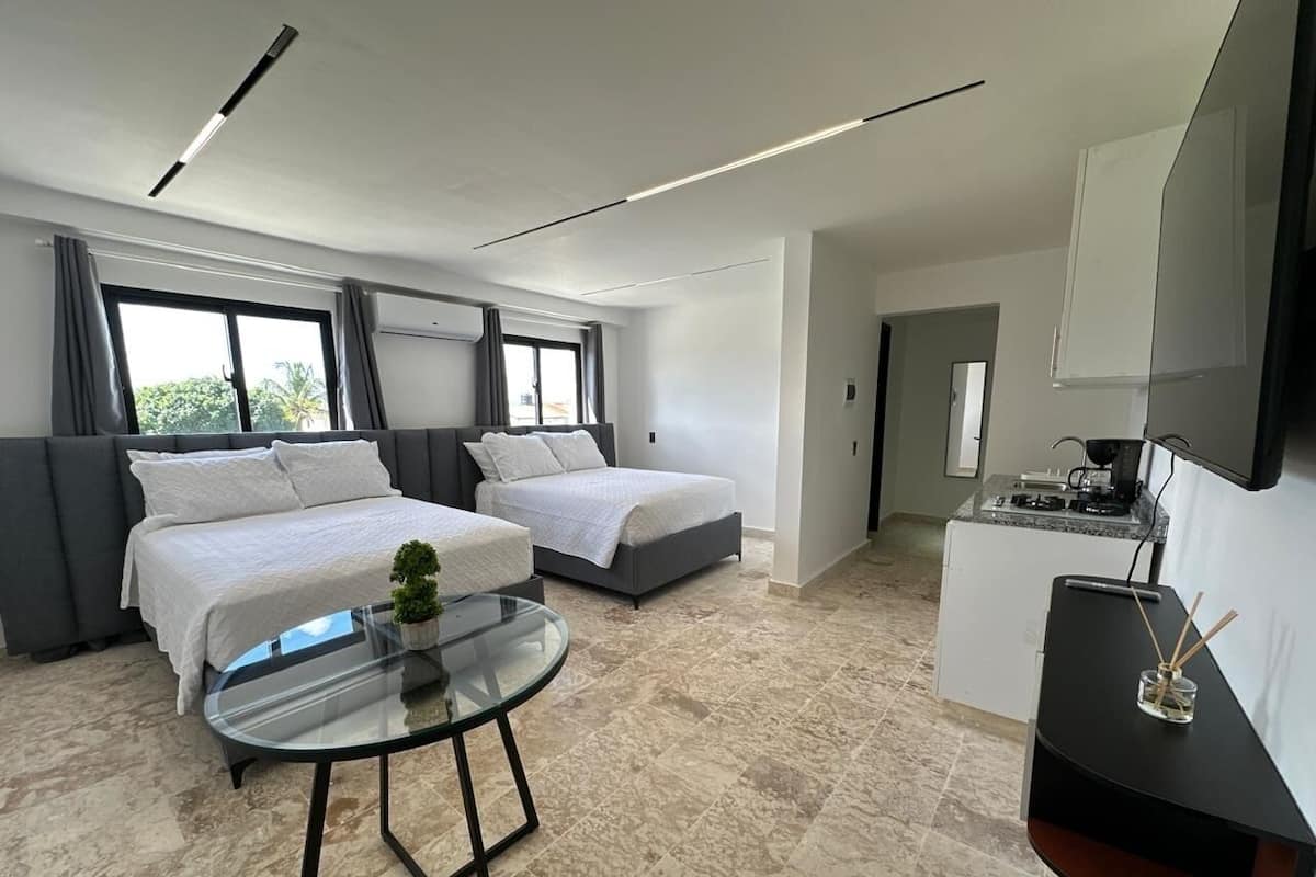 A modern hotel room with two beds, a glass coffee table, a small plant, a kitchen area, and a large window.