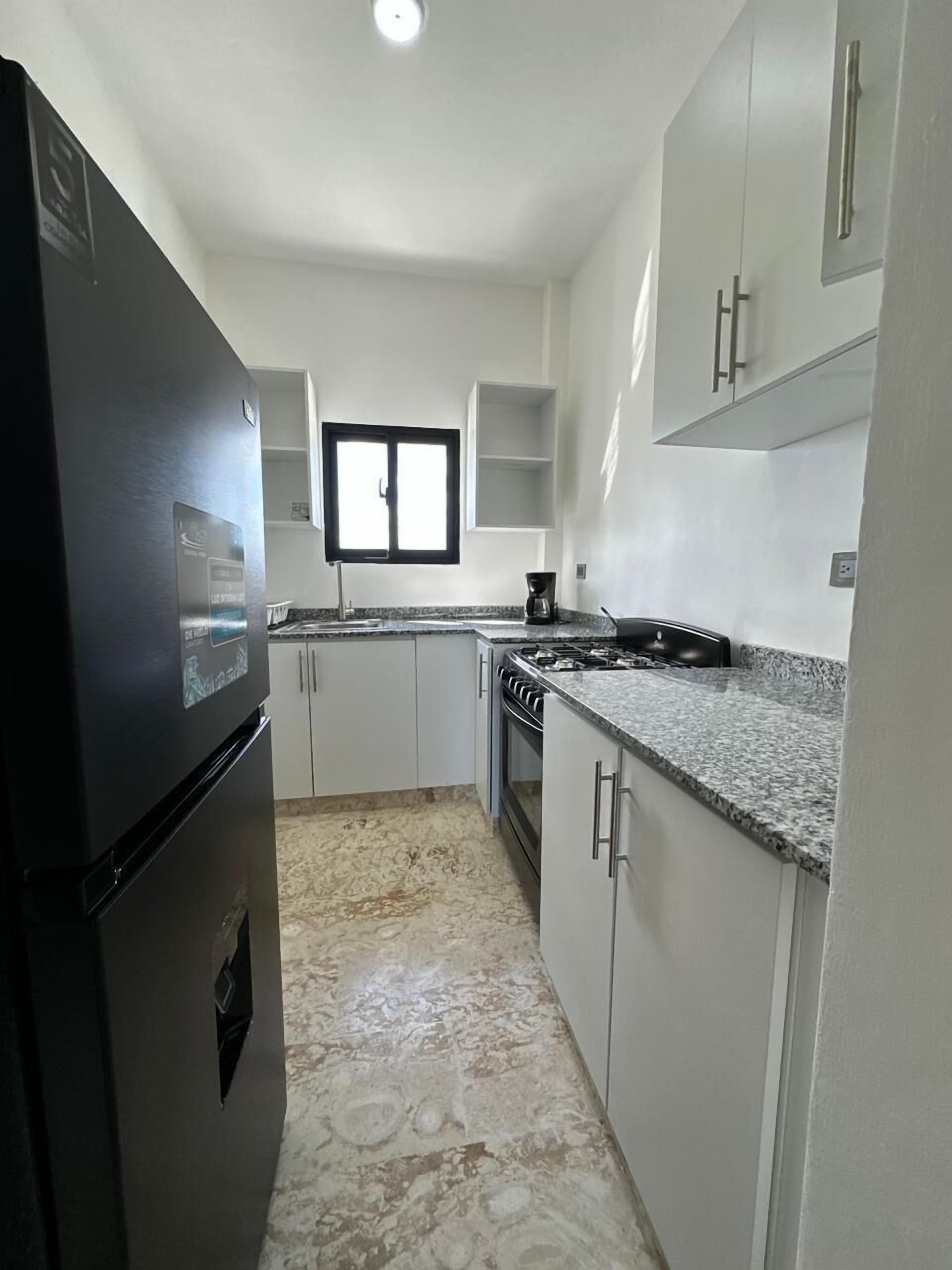 Une cuisine avec des meubles blancs, un réfrigérateur noir, une fenêtre et une cuisinière à gaz.