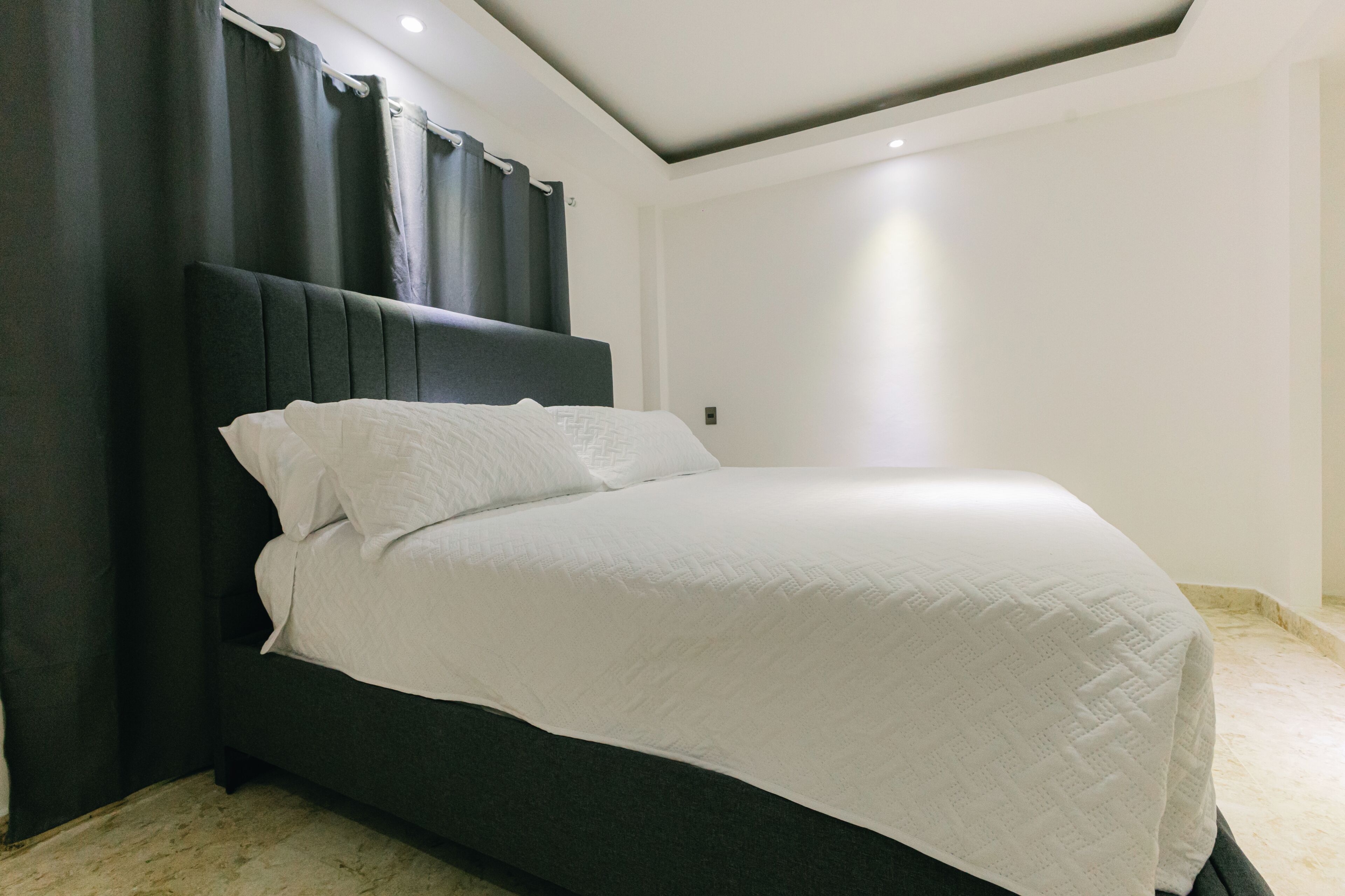 Una cama con cabecera oscura, ropa de cama blanca y una pared lisa con una luz en el techo.