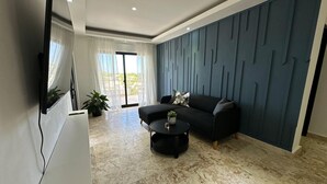En moderne stue med et fladskærms-tv, en sort sofa, et rundt sofabord og en væg med et mønster.