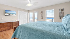 A bedroom with a bed, a ceiling fan, a TV, and a view of the ocean through the windows. - Sound-View Beach Retreat w/ Multiple Decks, Kayaks & Bikes  Island Vibes (Navarre)