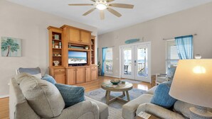 A spacious living room with a ceiling fan, wooden cabinets, and a large window with blue curtains. - Sound-View Beach Retreat w/ Multiple Decks, Kayaks & Bikes  Island Vibes (Navarre)