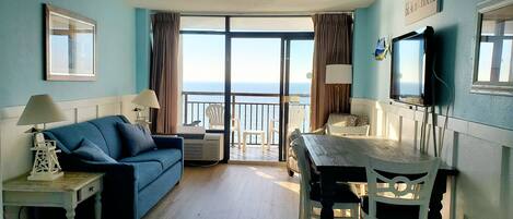 A room with a blue sofa, a dining table with chairs, a TV, and a view of the sea through large windows.