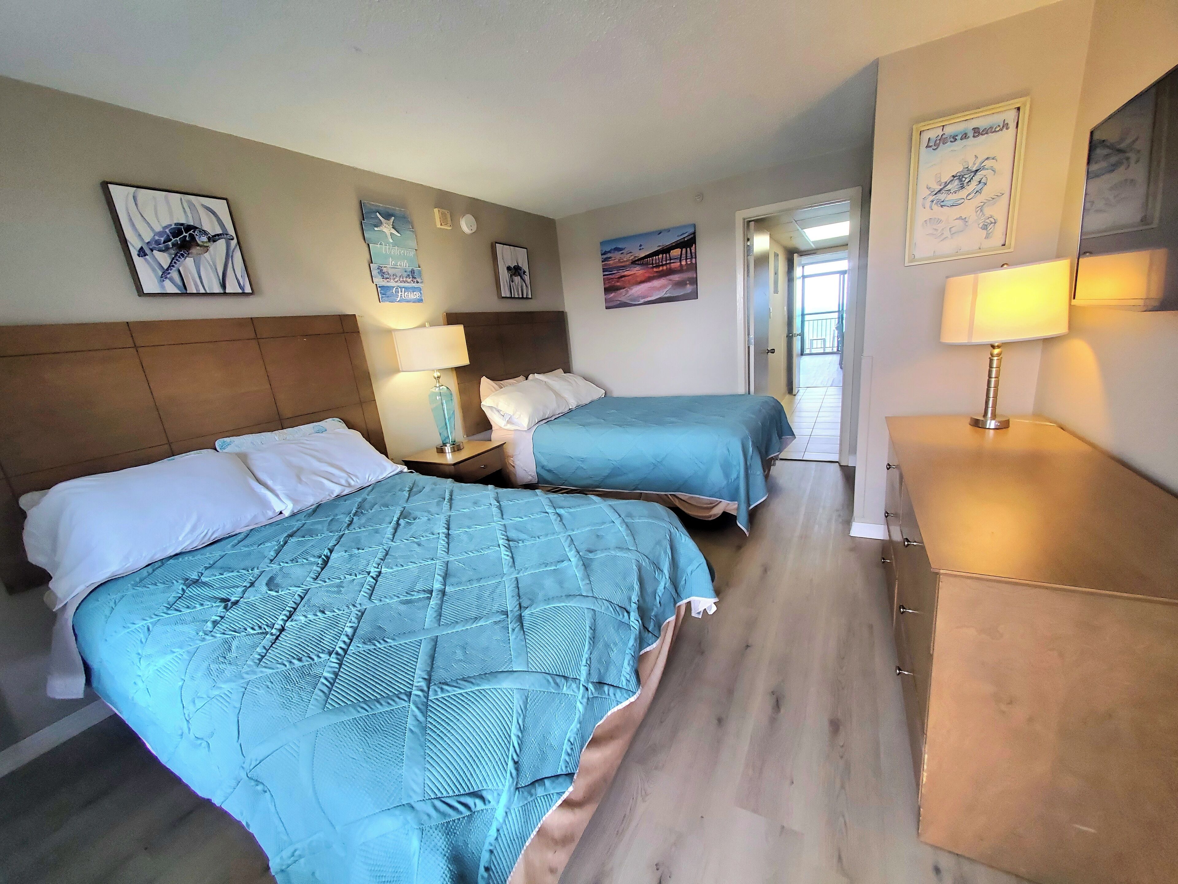 Quarto de hotel com duas camas, uma mesa de cabeceira com abajur e cômoda. Há quadros nas paredes e uma porta que dá para outro aposento.
