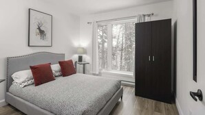 Ein Schlafzimmer mit einem Bett, einem Nachttisch, einem Fenster mit Blick auf Bäume und einem dunklen Kleiderschrank.