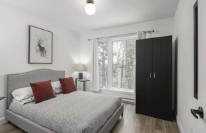 A bedroom with a bed, a nightstand, a window with a view of trees, and a dark wardrobe. - NatureX | Spa | Rivière | Dogfriendly (Matawinie Regional County Municipality)