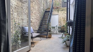 A narrow outdoor space with a stone wall, a metal staircase, a cushioned chair, a small table, and potted plants.