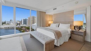 A modern hotel room with a large bed, a bench, and a nightstand. The room features a large window with a city view. - 1 BR Suite @ 1 Hotel | Gym+Pool+Rooftop (Miami Beach)