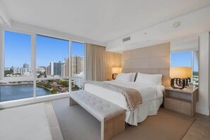Habitación de hotel moderna con una cama grande, un banco y una mesita de noche. La habitación cuenta con un ventanal con vistas a la ciudad.
