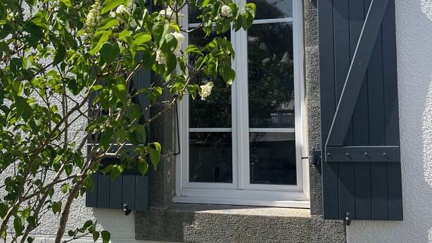 A building with a white textured exterior, a window with white shutters, and a tree with white flowers in front. - Crozon-Morgat: Renovated family home for 6-8 people 5 minutes from the beach (Crozon)
