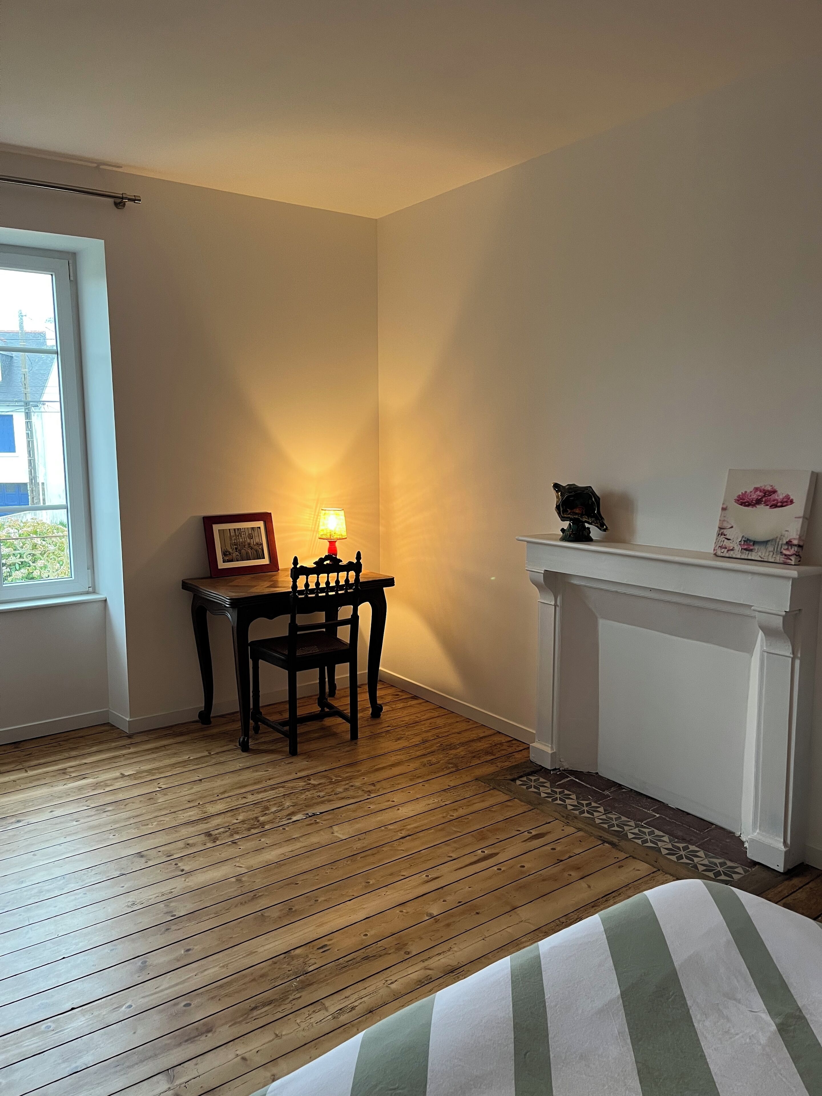 Une pièce avec un plancher en bois, une table à manger entourée de chaises, une lampe de table allumée, une cheminée avec un manteau et un tableau encadré accroché au mur.