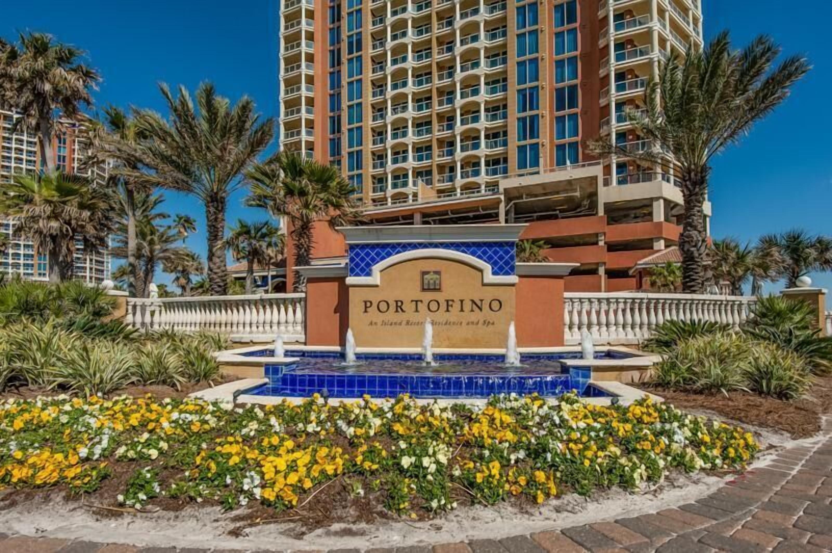 A hotel entrance with a sign reading "PORTOFINO," surrounded by palm trees and a flower bed.