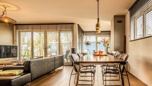 A modern living room with a large sofa, dining table, and chairs, large windows with blinds, and a television. - Nova Houseboat Deluxe + free bikes (Amsterdam)