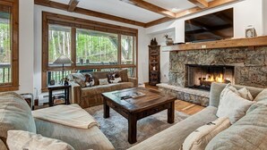 Ein gemütliches Wohnzimmer mit Kamin aus Stein, Holzbalken, großen Fenstern und bequemen Sitzgelegenheiten.