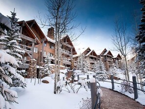 Lumipeitteinen mökki, jossa on useita puurakennuksia, kivipolku ja lumikuormaiset puut.