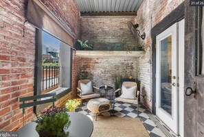 Een gezellig balkon met bakstenen muren, twee rieten stoelen, een klein rond tafeltje en een plant in pot.