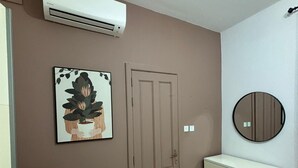 Una habitación con pared color beige, un equipo de aire acondicionado blanco, una puerta, un cuadro enmarcado, un espejo redondo y un aparador blanco.