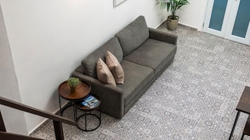 Un sofá gris con dos cojines, una mesita redonda con una planta y una revista, y un suelo estampado.