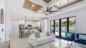 A modern living room with a white sofa, blue armchairs, and a glass coffee table. A kitchen with white cabinets and stainless steel appliances is visible in the background. - 1720 SW 19th PL (Cape Coral)