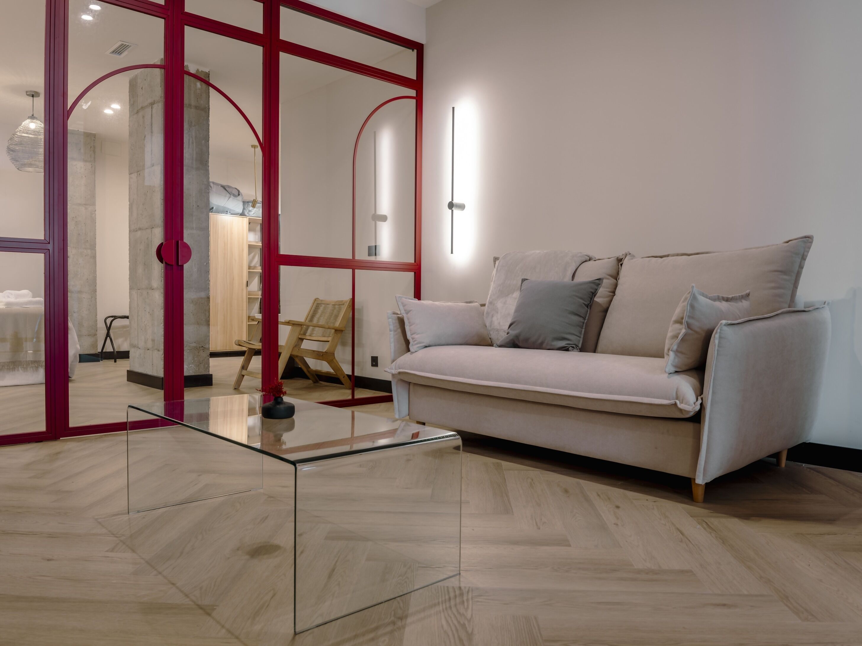 Un salon moderne avec une table basse en verre, un canapé beige agrémenté de coussins, une chaise en bois et une cloison en verre avec un cadre rouge.