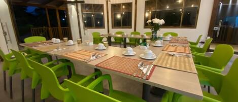 Comedor con una mesa larga dispuesta para varias personas, sillas verdes y grandes ventanales que dejan entrar luz natural.