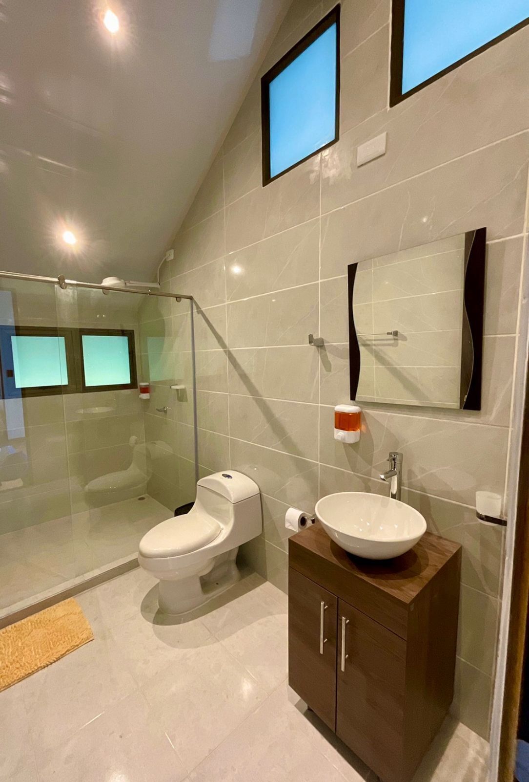 Un baño moderno con cabina de ducha de vidrio, inodoro, lavabo con tocador de madera y dos pantallas empotradas.