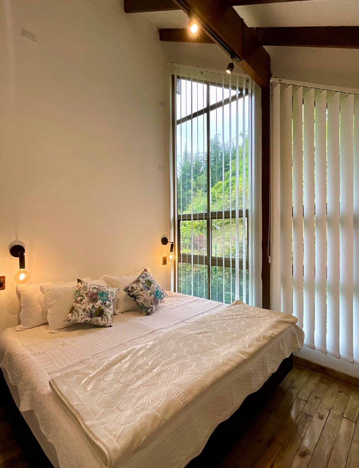 A bedroom with a large bed, a window with blinds, and a wooden floor.