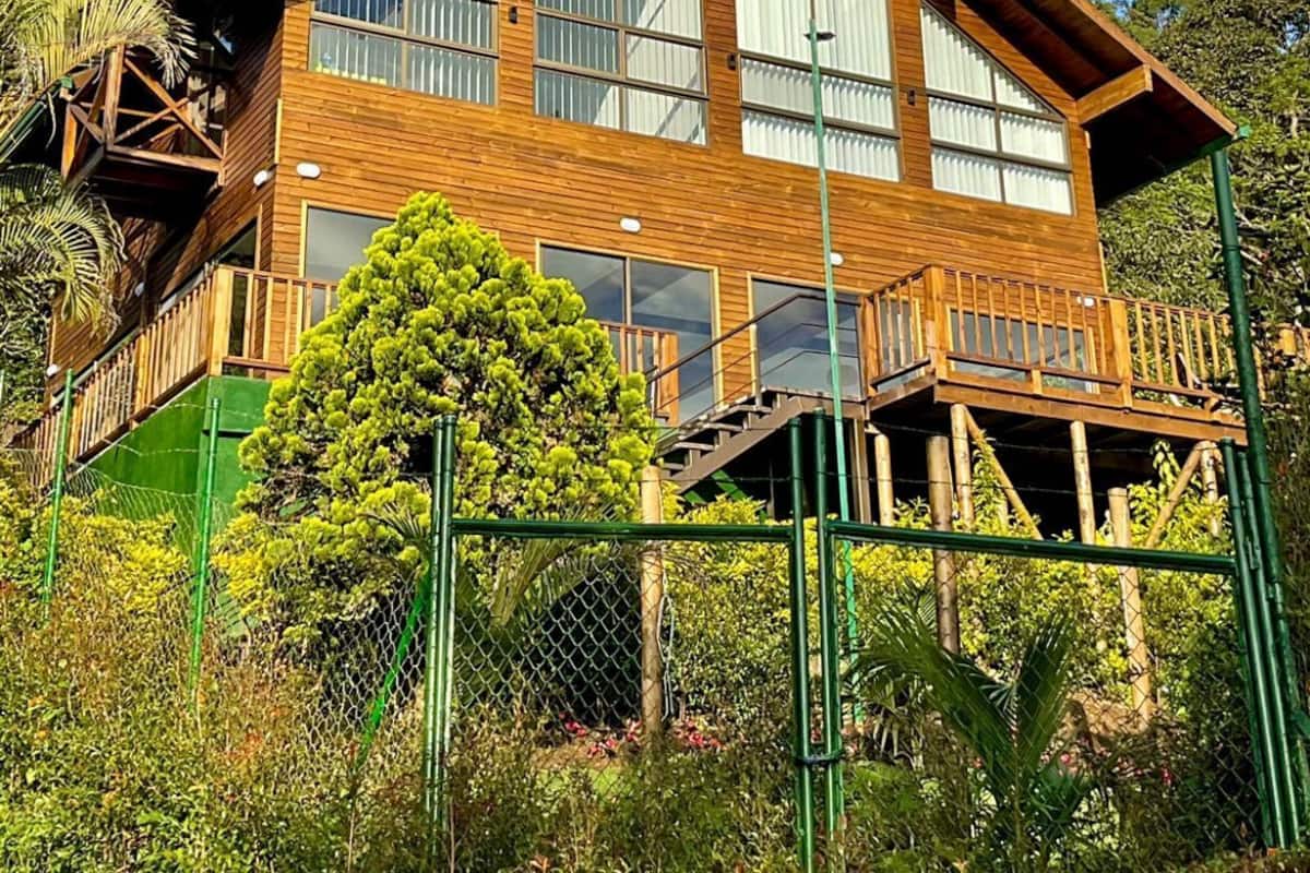 A two-story wooden house with large windows and a balcony, surrounded by greenery.