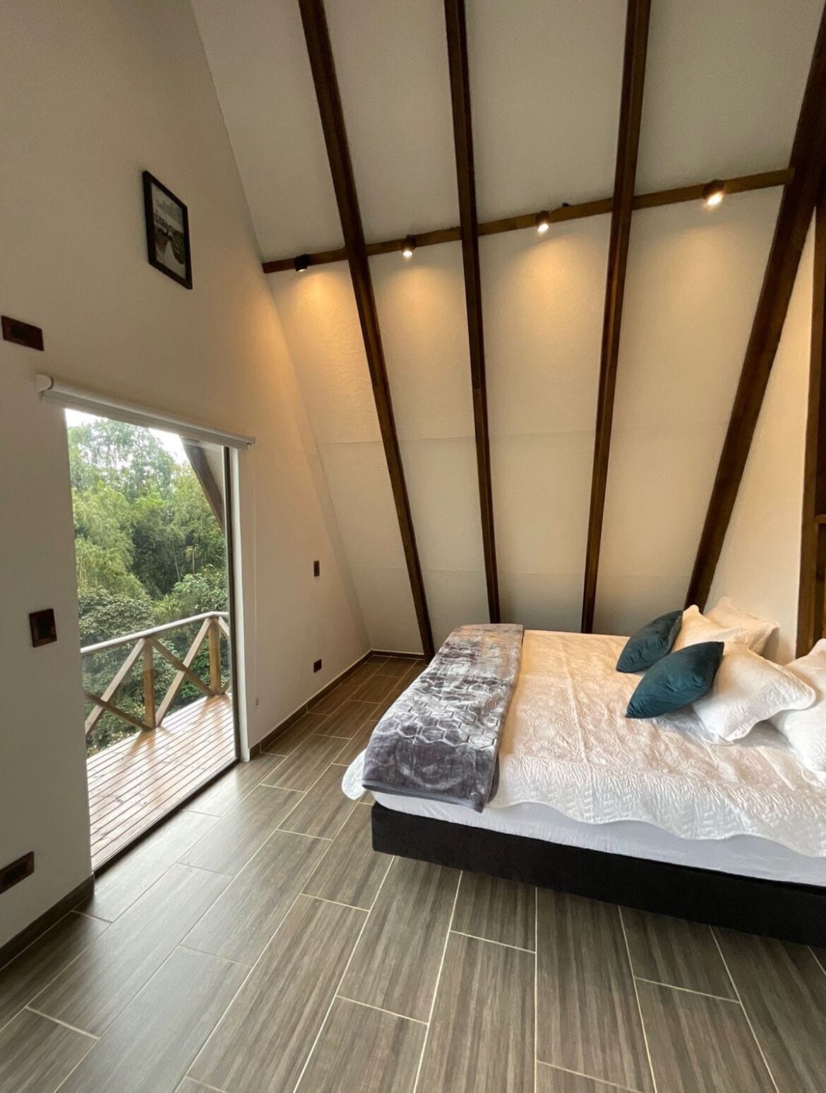 A bedroom with a large bed, a balcony with a wooden railing, and a view of greenery outside.