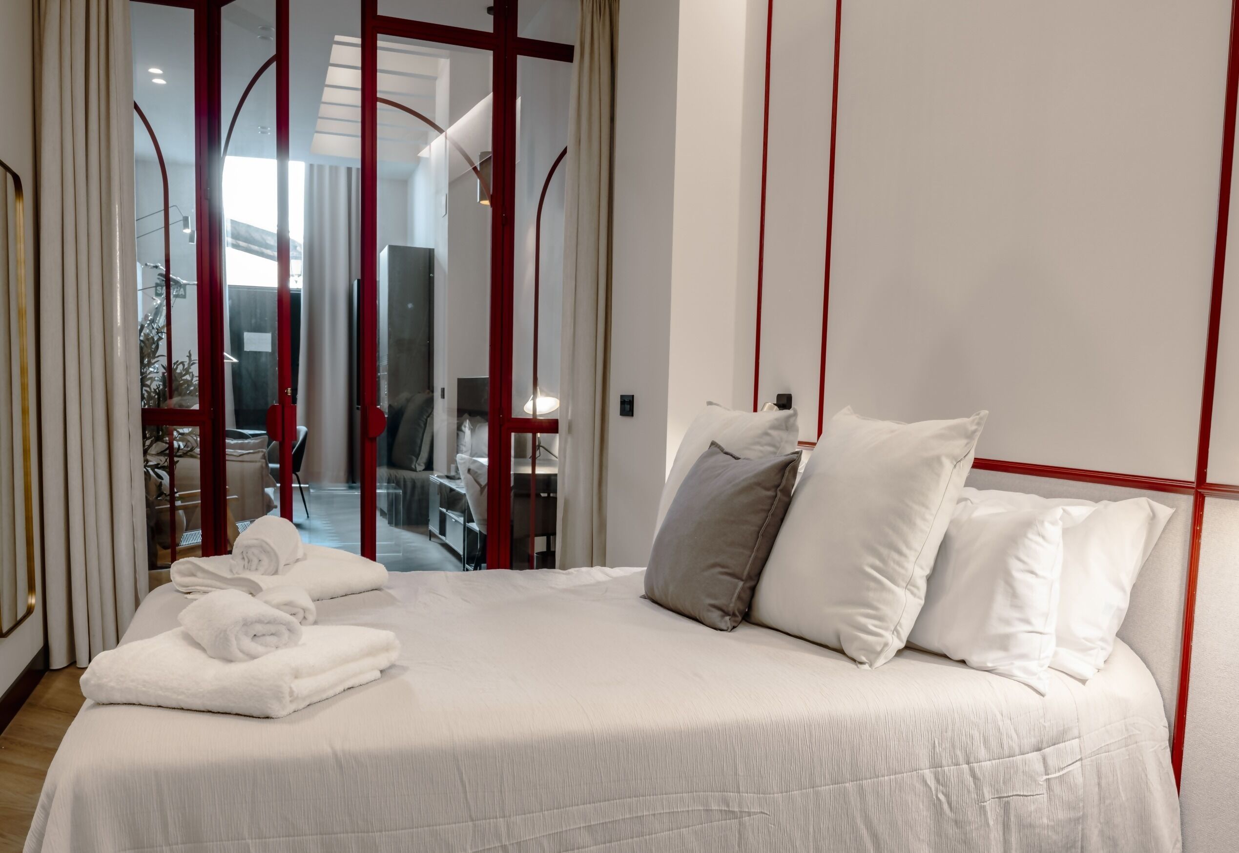 A neatly made bed with white linens and pillows, a wooden floor, and a red-framed sliding door leading to a balcony.