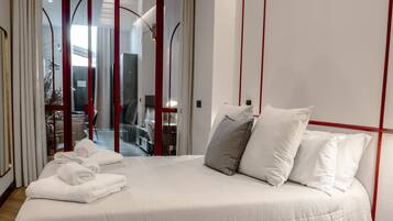 Una cama bien tendida con sábanas y almohadas blancas, suelo de madera y una puerta corredera con marco rojo que da a un balcón.
