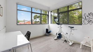 Une pièce spacieuse avec de grandes fenêtres, une table blanche, un vélo d’appartement et un petit tabouret blanc.