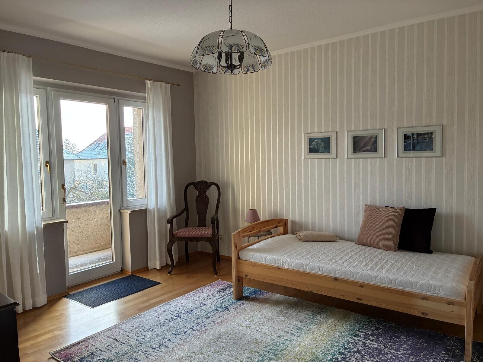 A bedroom with a bed, a chair, a hanging light fixture, and a view through a window.