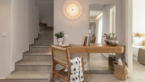 Een trap die leidt naar een tweede verdieping, een houten bureau met een plant, boeken en decoratieve objecten, en een bed met kussens in de aangrenzende kamer.