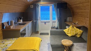 Ein gemütliches Blockhäuschen mit Küchenzeile, einem Sofa mit gelbem Kissen und Meerblick durch eine Tür.
