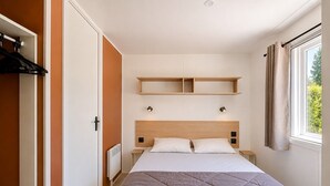 Ein Schlafzimmer mit Bett, Nachttisch, Ablage über dem Bett, Heizkörper und einem Fenster mit Vorhängen.