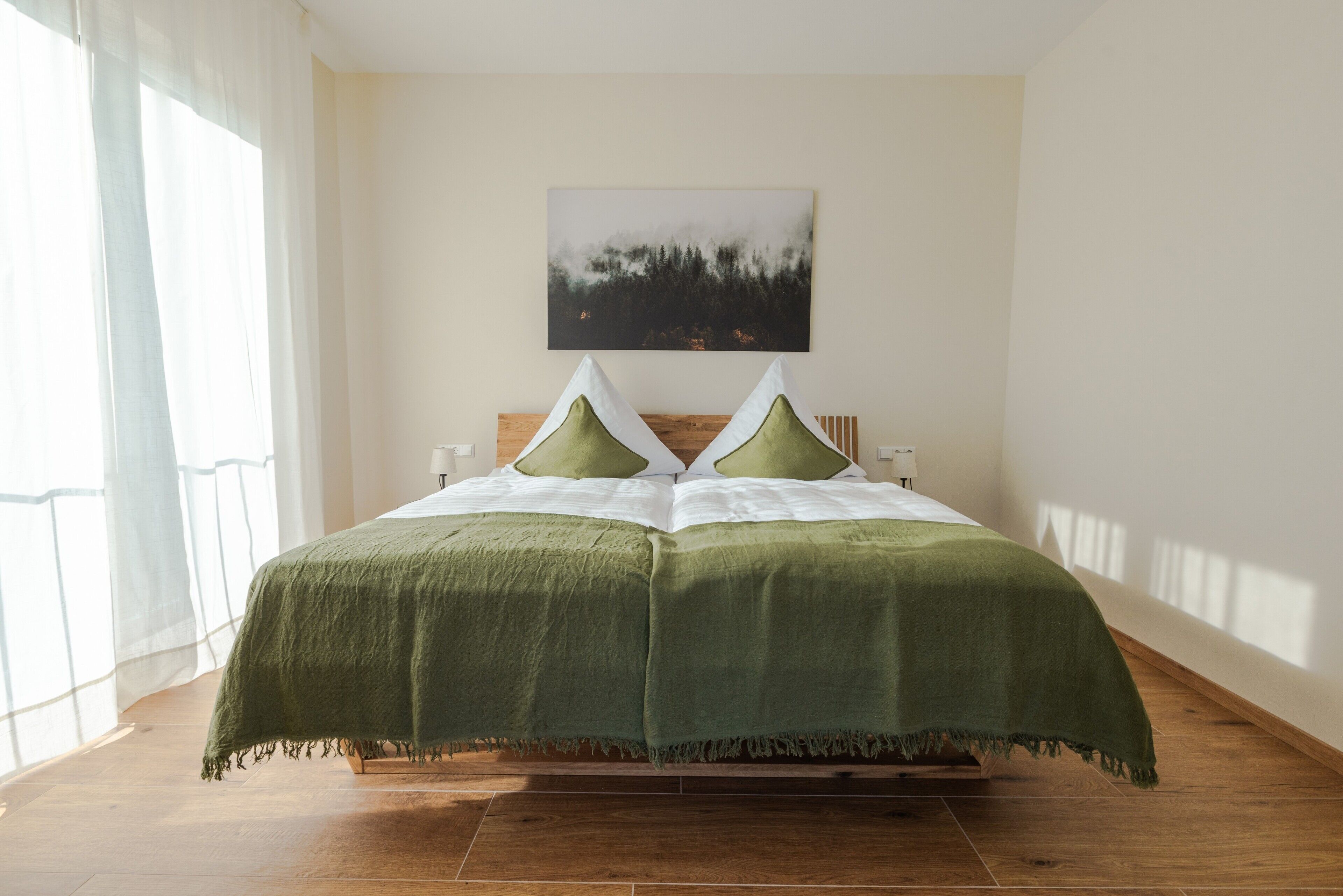 Uma cama com colcha e travesseiros verdes, cabeceira de madeira, mesa de cabeceira e um quadro de uma floresta na parede.
