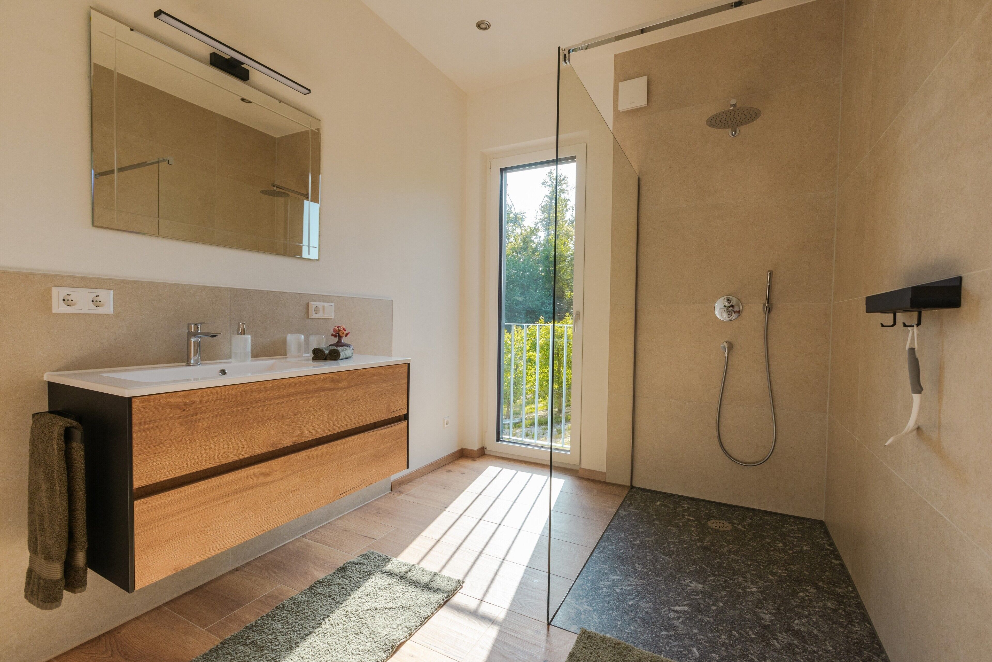 A modern bathroom with a wooden vanity, a large mirror, a shower with a glass door, and a view of greenery outside.