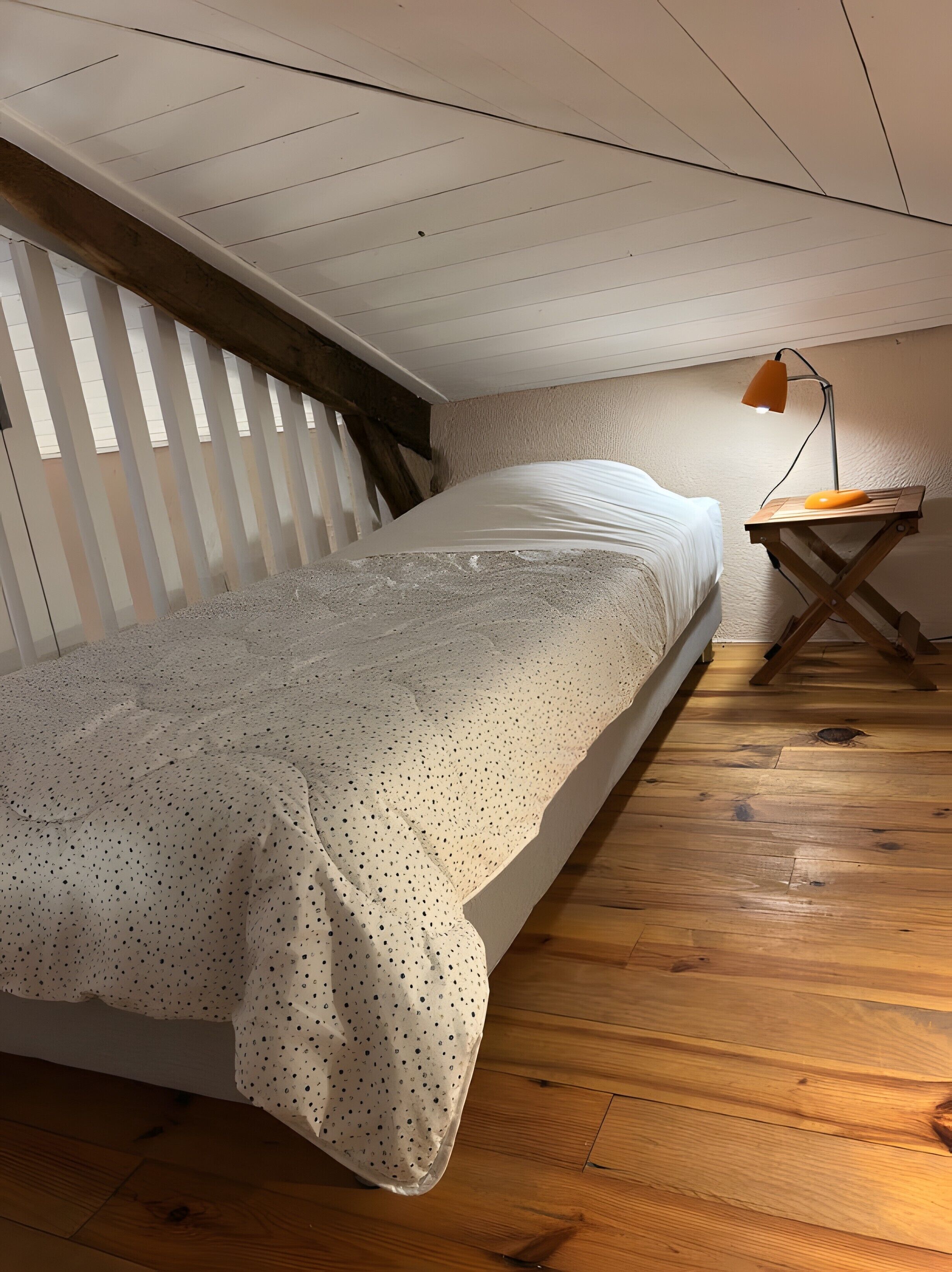 A bedroom with a bed, wooden floor, slanted ceiling, and a desk with a lamp.