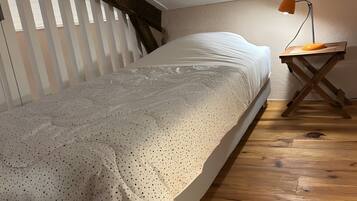 A bedroom with a bed, wooden floor, slanted ceiling, and a desk with a lamp.