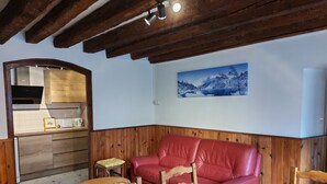 Un salon chaleureux avec un plafond en bois, un canapé en cuir rouge, une table à manger en bois et une cuisine visible à travers une porte ouverte.