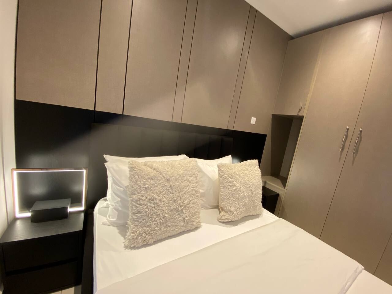 A modern bedroom with a bed, two fluffy white pillows, a bedside table with a lamp, and built-in wardrobes.