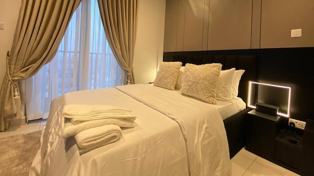 A neatly made bed with white linens and pillows, a bedside table with a lamp, and a view of the outside through large windows with beige curtains. - Unique Abodes (Accra)