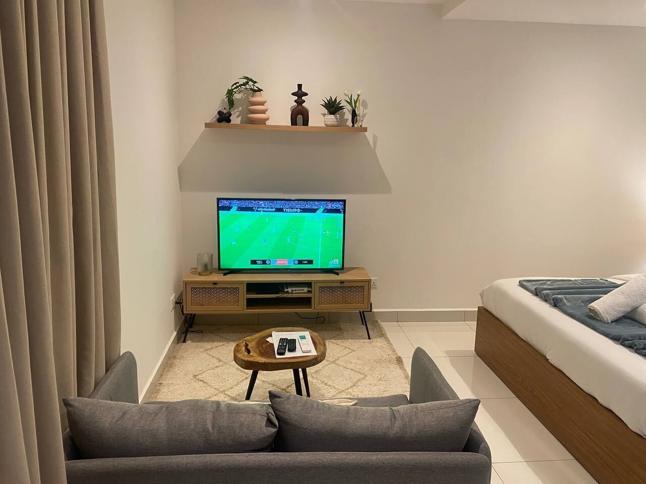 A modern living room with a flat-screen TV on a wooden stand, a grey sofa, a wooden coffee table, and a bed with blue bedding.