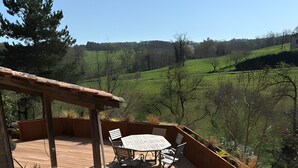 Una terraza de madera con una mesa y sillas, rodeada de macetas de piedra y vistas a un paisaje verde.